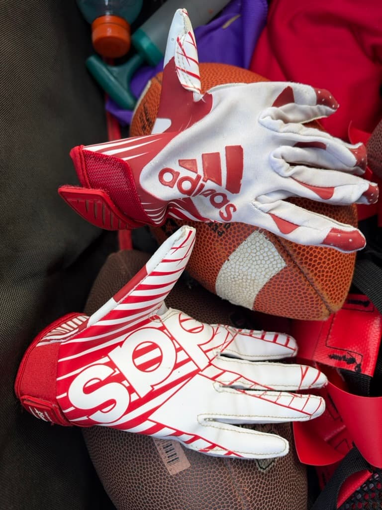 My son’s red and white Adidas receiving gloves resting on footballs in our gear bag.